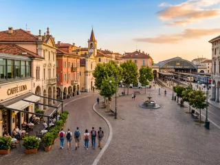 Sonnenbeschienener südeuropäischer Altstadtplatz im Abendlicht mit historischen Arkadengebäuden, Straßencafé, Kirchturm und einem Bahnhof mit Zügen rechts; junge Reisende mit Rucksäcken schlendern über das Kopfsteinpflaster